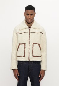 Veste en polaire crème avec un design à rayures brun. Dotée d'un large col, d'une fermeture éclair devant et de deux poches surdimensionnées avec des accents bruns.