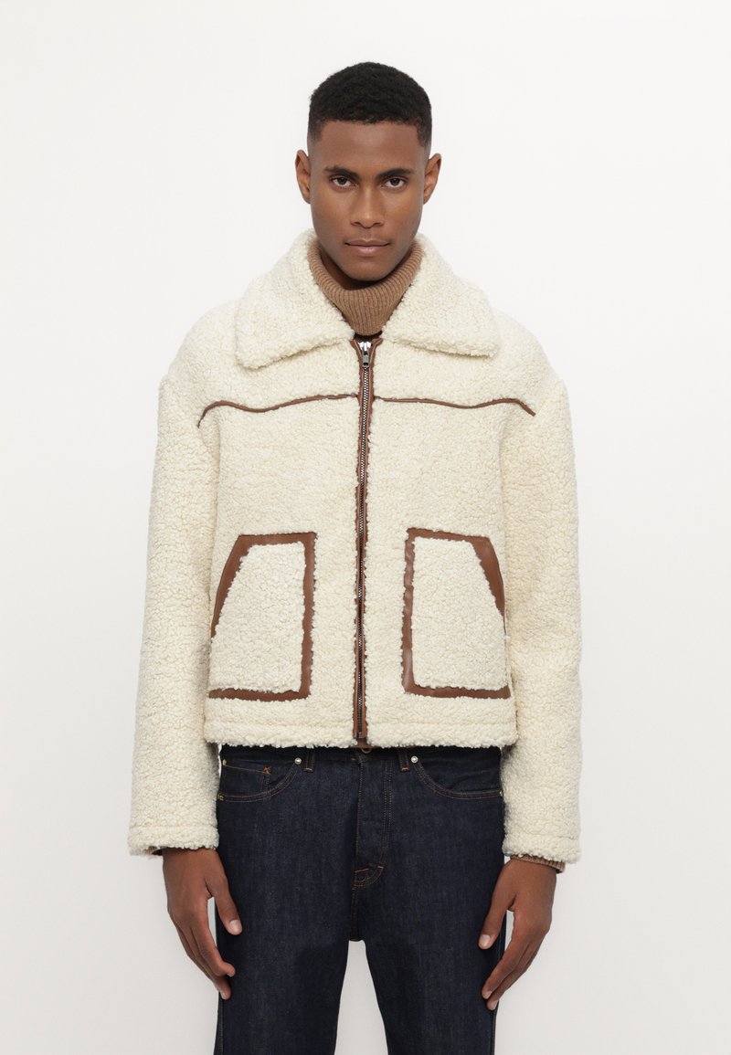 Fleece jacket in cream with a brown line design. Features a wide collar, front zip, and two oversized pockets with brown accents.