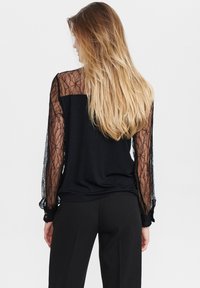 Femme avec de longs cheveux blonds portant un blouse noire avec des manches en dentelle florale transparente et un pantalon noir, vue de dos sur fond blanc.