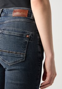 Vue arrière d'une personne portant un jean en denim bleu foncé avec une étiquette en cuir de la marque sur la ceinture et la main détendue le long du corps.