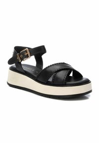 Sandalias de plataforma de piel negra con diseño de correas cruzadas, hebilla ajustable y costuras texturizadas. Suela blanca gruesa con fondo negro.