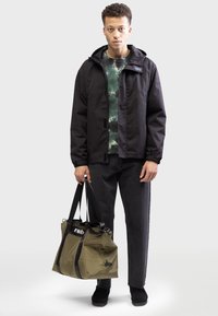 Zwarte waterdichte jas met capuchon, groen tie-dye shirt, donkere spijkerbroek en zwarte schoenen. Draagt een olijfgroene tote bag met zwarte banden.