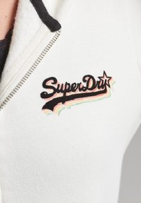 Sudadera blanca con cremallera y cuello negro. Presenta un colorido logo de "SuperDry" con franjas rosas, verdes y naranjas debajo del texto. Textura suave.
