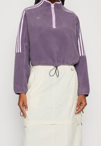 Lila fleece-tröja med halvmeterdragkedja, vita randiga detaljer på ärmarna och dragsko i nederkanten, bärs över en krämfärgad kjol.