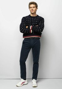 Navy knitted sweater with white speckles, red and cream striped cuffs, paired with dark pants and white sneakers with red and blue accents.