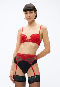 Červená čipkovaná podprsenka s kostícami a nastaviteľnými ramienkami, v kombinácii s čiernymi nohavičkami s vysokým pásom, ktoré majú červené čipkové detaily a podväzkové pásy.