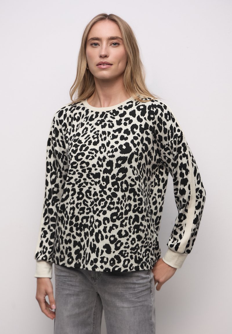 Frau mit langem blondem Haar, die ein schwarz-weißes, langärmliges Hemd mit Leopardenmuster und graue Jeans trägt, steht vor einem einfarbigen Hintergrund.