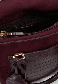 Burgunderfarvet taske med ruskindsmateriale, en lynlåslomme indeni og en tekstureret læderdetalje med guldlåg.