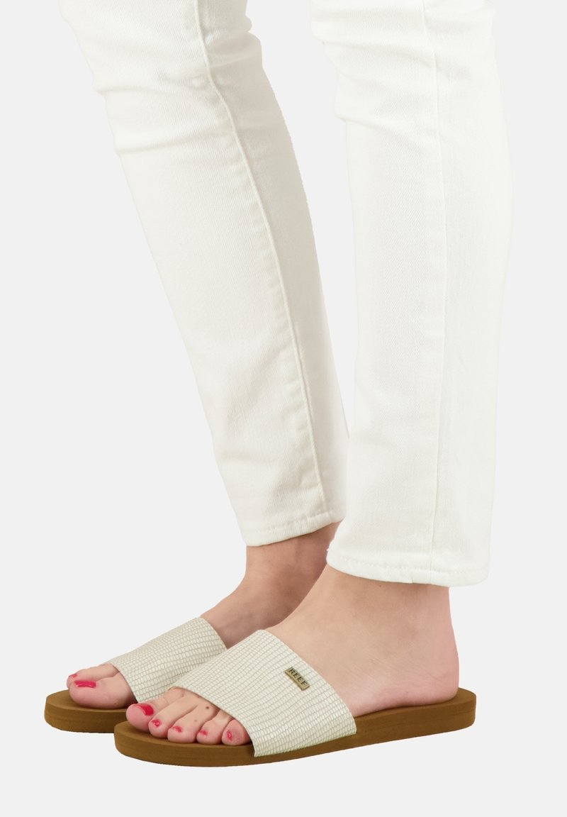 Des sandales à glissière beiges avec une lanière texturée et une semelle en caoutchouc marron, associées à un jean skinny blanc. Les ongles des orteils sont peints en rose.