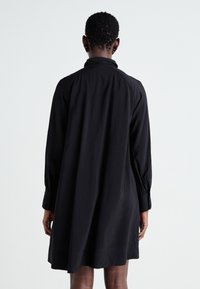Robe noire à manches longues, avec un poignet boutonné et une coupe décontractée. Fabriquée en tissu lisse avec un détail de col et une finition soignée.