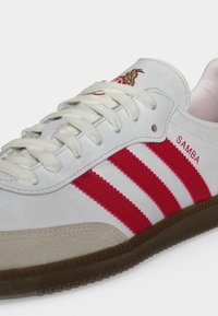 Λευκό αθλητικό παπούτσι Adidas Samba με κόκκινες ρίγες, λευκά κορδόνια, μπεζ σουέντ μυτερό μέρος και καφέ καουτσούκ σόλα, απεικονίζεται σε γωνιακή κοντινή λήψη.