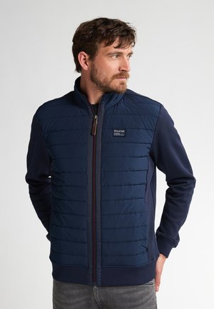 Chaquetón navy que presenta un frontales acolchado con costuras forradas, cremallera completa y tela contrastante en las mangas y los laterales. Parche con el logo en el pecho.
