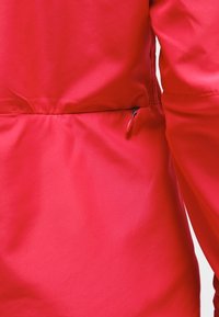 Acercamiento de una chaqueta roja que muestra un tejido suave y un detalle de un bolsillo con cremallera horizontal cerca de la cintura, en el lado derecho.