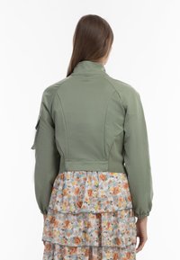 Olivengrøn cropped jakke med stiv krave, elastiske manchetter og et struktureret panel; lagdelt blomsterstof med orange og gule nuancer nedenfor.