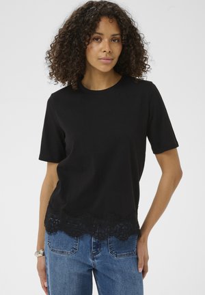 Femme aux cheveux bouclés portant un haut noir à manches courtes avec un ourlet en dentelle et un jean en denim bleu, posant devant un fond clair uni.