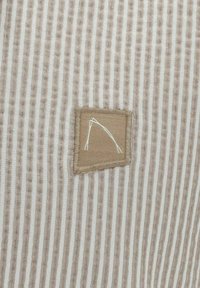 Tejido con patrón de rayas verticales en blanco y beige claro; presenta una etiqueta cuadrada de color beige cosida con bordado blanco.