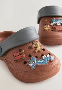 Sabots en caoutchouc marron avec une sangle grise, présentant des détails des personnages Tom et Jerry et des trous de ventilation pour la respirabilité.