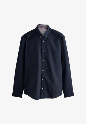 Tamsiai mėlyna medvilninė marškinėliai mygtukais, turi languotą raštą ant apykaklės viduje ir mažą raudonos spalvos karūnos logotipą ant krūtinės.