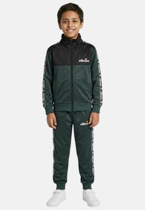 Garçon souriant debout, portant un survêtement Ellesse vert foncé et noir avec des bandes à logo sur les manches et le pantalon, assorti à des baskets blanches.