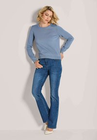 Frau, die an einer Wand lehnt, trägt einen hellblauen Pullover, blaue Jeans und weiße spitze Schuhe, schaut nach unten mit einer Hand auf der Hüfte.