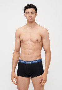 Zwarte boxershorts met een blauwe elastische tailleband waarop "HUGO" staat. De stof lijkt glad, met een strakke pasvorm en zonder zichtbare patronen.