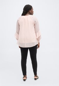 Γυναίκα με πλεξούδες, ντυμένη με διάφανη ροζ μπλούζα με βολάν στα μανίκια, μαύρο παντελόνι και μαύρα σανδάλια, όρθια με την πλάτη στραμμένη.