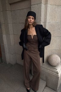 Bruine jumpsuit met een diepe halslijn, zwarte oversized jas, zwarte handschoenen en een zwarte hoed; omringd door stenen architectuur.