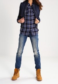 Person wearing a navy jacket over blue and white plaid shirt, faded blue jeans, and tan lace-up boots standing with hands adjusting jacket.