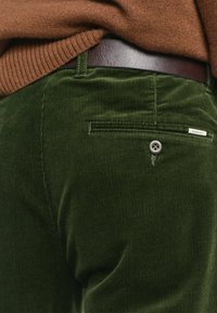 Grüne Cordhosen mit vertikalen Rippungen, ausgestattet mit einer Gesäßtasche, Knopfdetails und einem dezenten Logosticker. Kombiniert mit einem braunen Pullover und Gürtel.