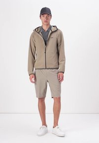 Lichtgewicht beige jas met een zwarte rits en afwerking, gecombineerd met bijpassende shorts. Model draagt een donkere pet en witte sneakers. Eenvoudig ontwerp.