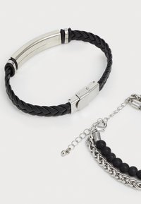 Brățară din piele împletită neagră cu închizătoare din metal de culoare argintie, asociată cu o brățară din mărgele și lanț, având mărgele negre și accente metalice.