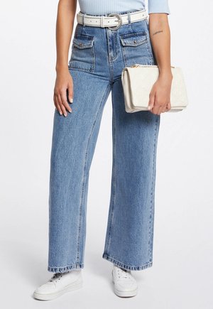 Frau trägt hellblaue Jeans mit großen vorderen Taschen, weißen Gürtel, weiße Turnschuhe, hält eine weiße gesteppte Handtasche vor weißem Hintergrund.