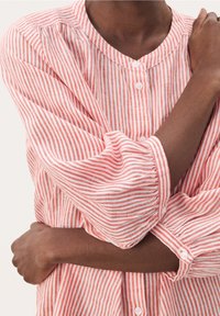 Camisa de botones a rayas rojas y blancas con cuello redondo. La tela parece ser texturada, con mangas largas abullonadas y puños abotonados.