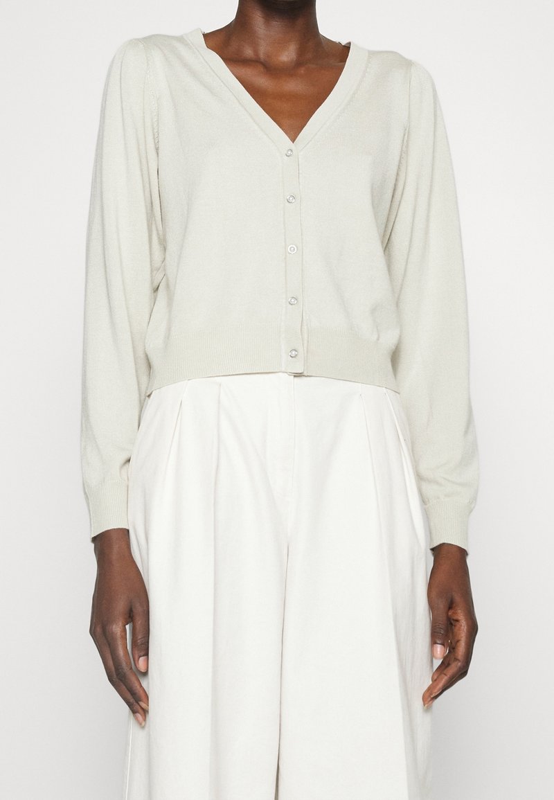 Personne portant un cardigan beige clair boutonné avec manches longues et un pantalon blanc plissé ample à taille haute sur un fond uni.