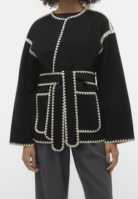 Schwarze Jacke mit langen Ärmeln, die über einen kontrastierenden weißen Wellenrand und eine taillierte Taille verfügt. Beinhaltet Taschen und hat einen lässigen Schnitt.