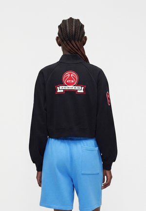Person med fletninger iført en sort Jordan jakke med rød logo og blå shorts, står med ryggen mod en ensfarvet baggrund.