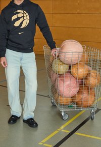 Sudadera negra con un gráfico de baloncesto dorado, jeans de mezclilla azul claro y zapatos negros al lado de una canasta de alambre llena de balones de baloncesto de varios colores.