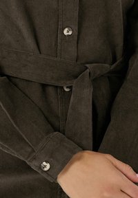 Braunes Cordhemd mit Knöpfen, einem gebundenen Gürtel und langen Ärmeln. Der Stoff hat eine gerippte Textur und einen subtilen Glanz.