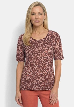Blondine vrouw met een kort mouwig, bruin-roze patroon top en roestkleurige broek, staand tegen een effen lichte achtergrond.