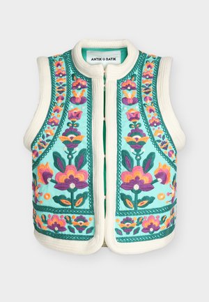 Mujer con blusa blanca con puños de encaje, chaleco floral bordado, vaqueros de pierna ancha, zapatos color burdeos y sosteniendo un bolso de mano burdeos.