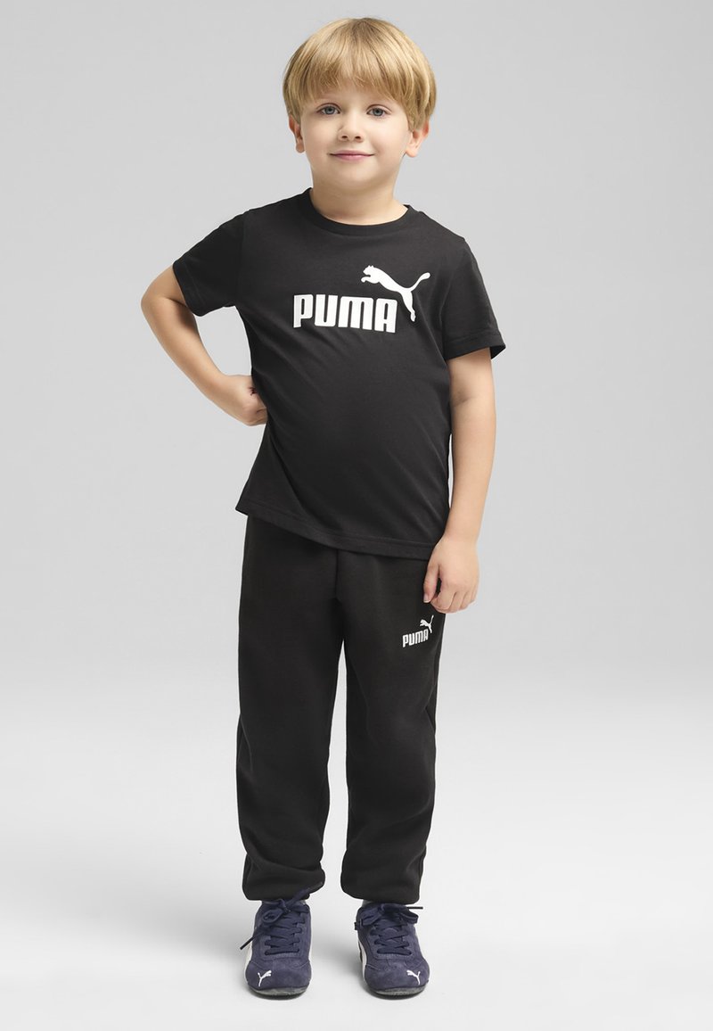 Juodas medvilninis marškinėlis su baltu "PUMA" logotipu, derinamas su juodomis sportinėmis kelnėmis, ant kurių yra mažas logotipas. Mėlyni sportiniai batai užbaigia aprangą.