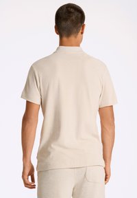 Beige kortärmad polo skjorta tillverkad av strukturerat tyg, med en nedvikt krage och en avslappnad passform, visad bakifrån.