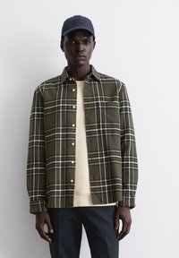 Camicia a quadri verdi con righe bianche e azzurre, con design a bottoni. Indossata sopra una camicia beige e abbinata a pantaloni scuri.