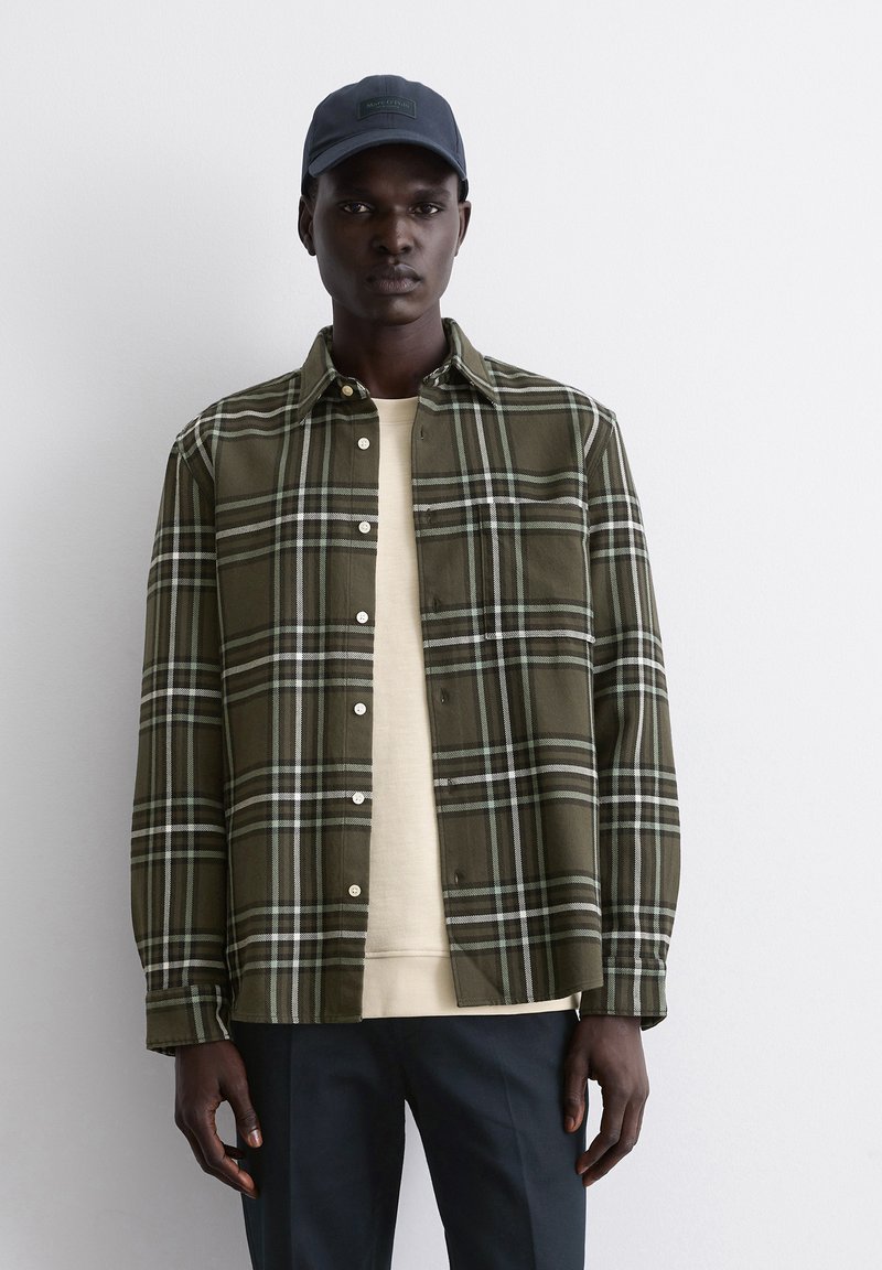 Camicia a quadri verdi con righe bianche e azzurre, con design a bottoni. Indossata sopra una camicia beige e abbinata a pantaloni scuri.