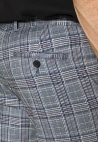 Gråblå rutiga byxor med klassisk passform, framfickor och svarta knappar, tillverkade av ett texturerat vävt material.