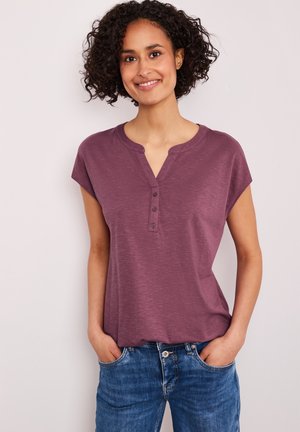 Kobieta o kręconych włosach, ubrana w fioletową koszulkę henley z krótkim rękawem i niebieskie dżinsy, uśmiechająca się z rękami w kieszeniach na jasnym tle.