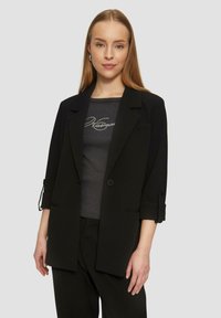 Schwarzes Blazer mit hochgerollten Ärmeln, Ein-Knopf-Verschluss und figurbetonter Passform, getragen über einem dunkelgrauen Oberteil mit silbernem Logo-Druck.