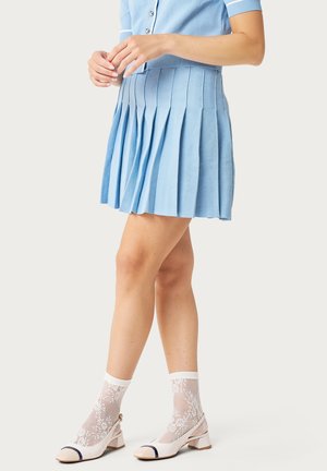 Mujer con falda plisada azul claro, top de manga corta azul a juego, calcetines blancos de encaje y zapatos beige de tacón bajo con punteras negras.