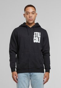 Svart hettegenser med glidelås, laget av bomull, med en frontlomme og hvit "STILL COLD" grafikk på venstre bryst. Løs passform med snor i hetten.