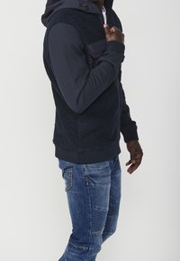 Mörkblå hoodiejacka med pälsstruktur på framsidan, släta ärmar och en framficka; bärs med ljusblå slitna jeans.